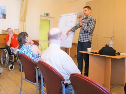 Prowadzący zajęcia przy tablicy, słuchacze na krzesłach i wózku
