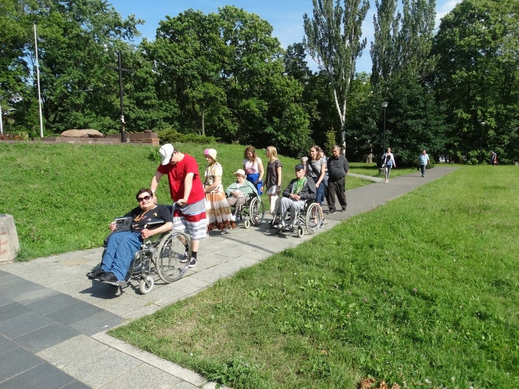Grupa mieszkańców, część na wózkach, spaceruje ścieżką w parku.