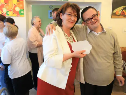 Kobieta w czerwonej sukience obejmuje mieszkańca w okularach, oboje trzymają opłatek