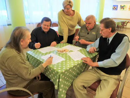 Mieszkańcy grają w bingo przy stole w grochy, opiekunka pochyla się nad nimi