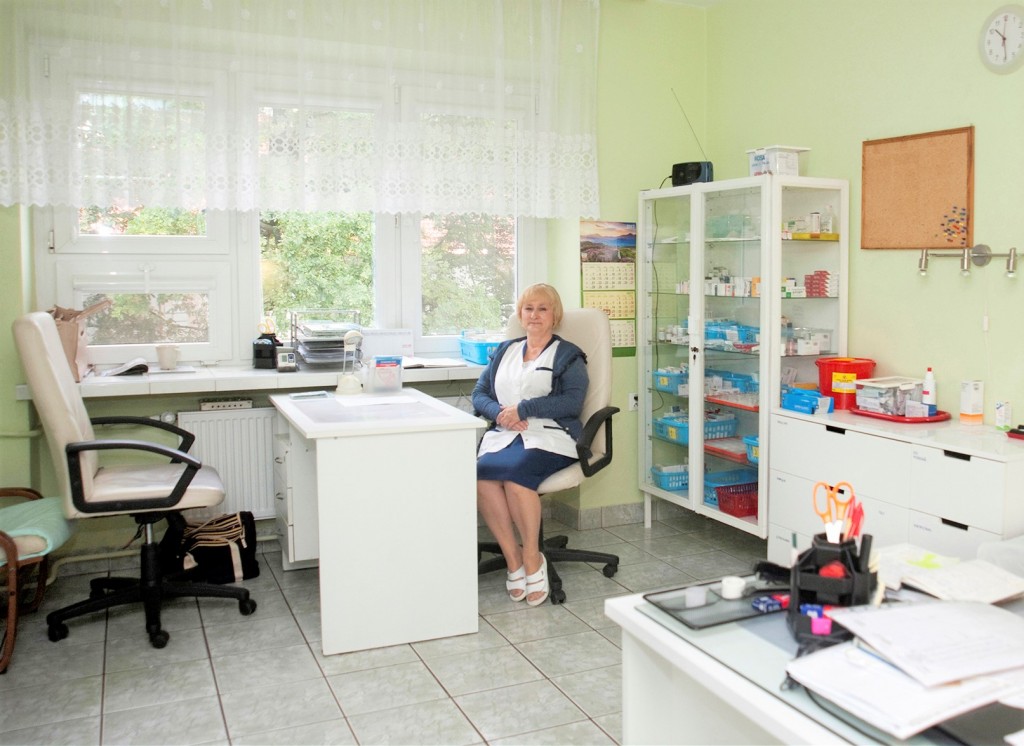 Gabinet pielęgniarski: pielęgniarka siedzi przy biurku, obok przeszklona szafka z lekami.