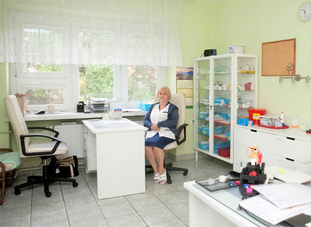 Gabinet pielęgniarski: opiekunka siedzi przy biurku, obok przeszklona szafka z lekami.