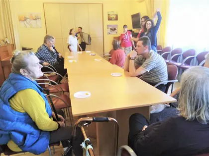 Mieszkańcy siedzą przy długim stole, w głębi akordeonista
