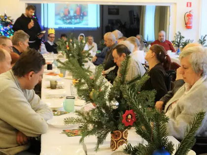 Mieszkańcy przy wigilijnym stole ze stroikami, ksiądz mówi do mikrofonu