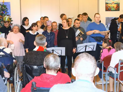Grupa kolędników śpiewa przy pulpitach przed mieszkańcami