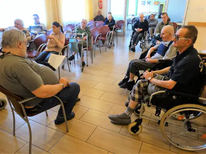 Mieszkańcy na wózkach i krzesłach słuchają czytania w świetlicy
