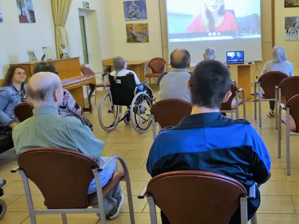 Mieszkańcy oglądają na ekranie warsztaty online prowadzone przez kobietę