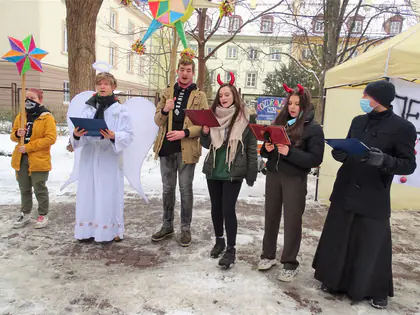 Kolędnicy jako aniołowie, diabły i ksiądz śpiewają z teczkami w dłoniach