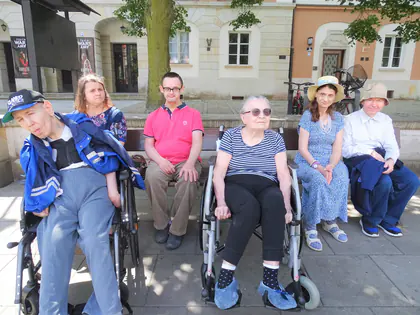 Mieszkańcy, część na wózkach, siedzą na ławeczkach na Starym Mieście