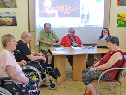 Mieszkańcy czytają teksty przy stole przed ekranem w świetlicy, słuchacze siedzą na wózkach