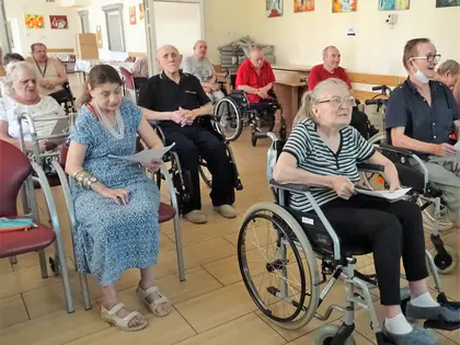 Mieszkańcy na wózkach i krzesłach śpiewają z kartkami w sali