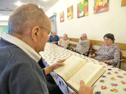 Mieszkaniec czyta książkę na głos przy stole, słuchają go mieszkanki