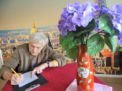 Mieszkanka pisze list przy stole z wazonem fioletowych kwiatów