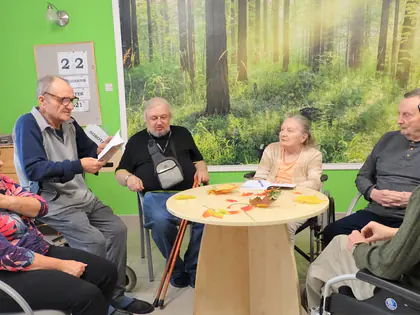 Mieszkańcy siedzą przy okrągłym stole z jesiennymi liśćmi i wspólnie piszą opowiadanie