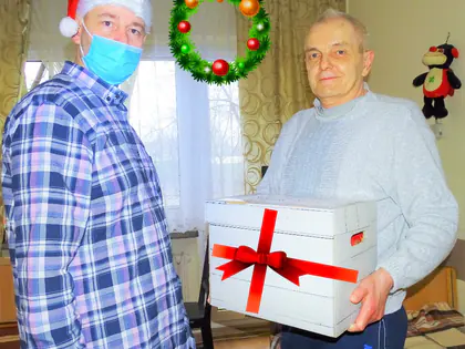 Mężczyzna w czapce Mikołaja i maseczce wręcza mieszkańcowi pudełko z kokardą
