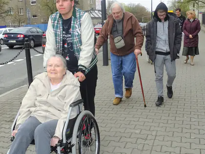 Młody mężczyzna pcha wózek z mieszkanką na chodniku, za nimi idą mieszkańcy i młodzież