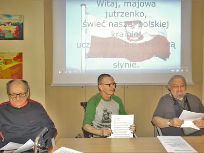 Mieszkańcy z kartkami siedzą przed ekranem z tekstem pieśni Witaj, majowa jutrzenko