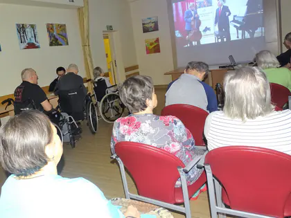 Mieszkańcy, część na wózkach, oglądają koncert wyświetlany na ekranie w sali