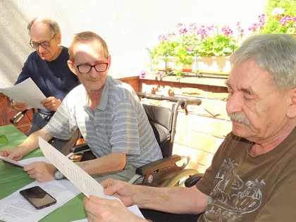 Mieszkańcy czytają teksty przy stole na tarasie z pelargoniami