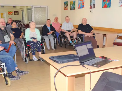 Mieszkańcy, część na wózkach, słuchają wykładu prowadzonego przy laptopie