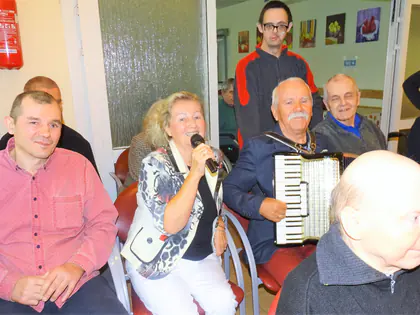 Kobieta śpiewa do mikrofonu, obok mężczyzna gra na akordeonie, wokół siedzą mieszkańcy
