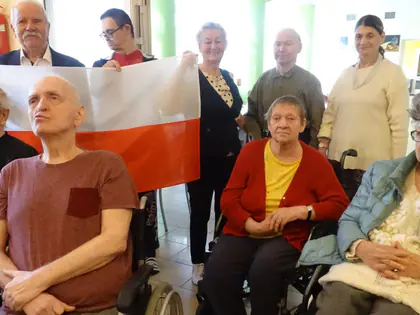 Mieszkańcy, część na wózkach, trzymają biało-czerwoną flagę podczas wspólnego śpiewania hymnu