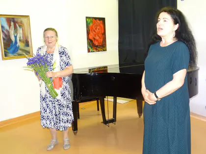 Pianistka stoi przy fortepianie obok kobiety trzymającej bukiet fioletowych kwiatów