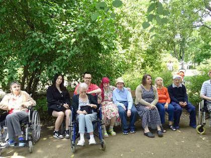 Mieszkańcy i opiekunowie siedzą na ławkach w zielonym parku