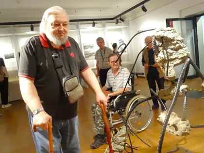 Mieszkańcy, jeden o lasce, drugi na wózku, zwiedzają wystawę w muzeum przy instalacji z metalowych prętów