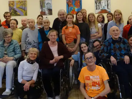 Grupa młodzieży i mieszkańców, część na wózkach, pozuje do wspólnego zdjęcia w sali z obrazami