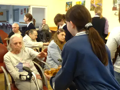 Uczennica z koszyczkiem podchodzi do starszej mieszkanki z balkonikiem