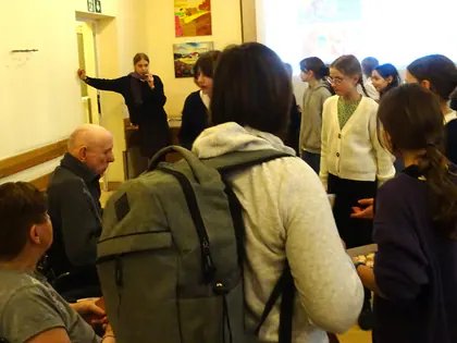 Uczennice rozmawiają z mieszkańcami i częstują ich, w tle drzwi i obrazy na ścianie