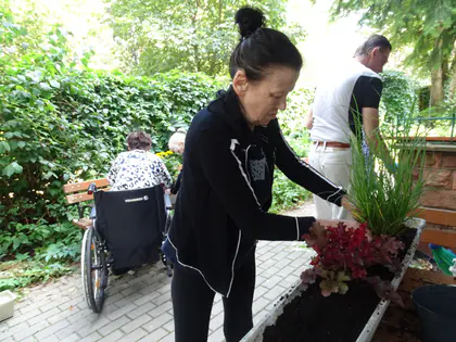 Kobieta sadzi rośliny w skrzynce, w tle mieszkanka na wózku i mężczyzna