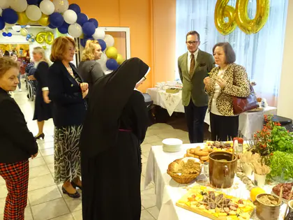 Goście przy stole z poczęstunkiem w sali ozdobionej balonami z liczbą 60, wśród nich siostra zakonna