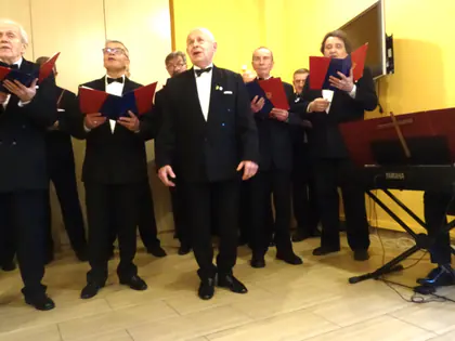 Chórzyści w frakach stoją w rzędzie, z boku pianista przy keyboardzie