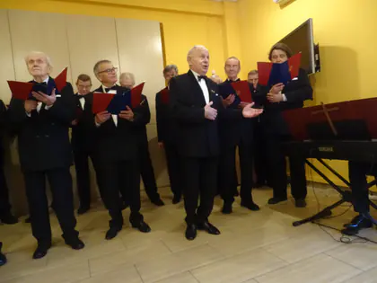 Chór męski w czarnych frakach z czerwono-granatowymi teczkami nutowymi śpiewa na tle żółtej ściany