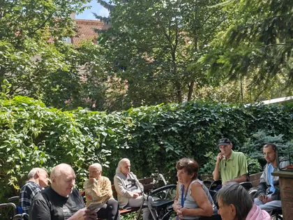 Mieszkańcy siedzą w ogrodzie wśród zieleni, część na wózkach