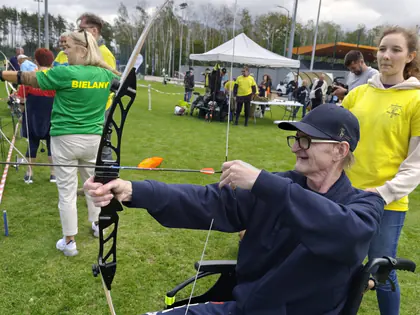 Mieszkaniec na wózku napina łuk na zawodach sportowych, obok stoją wolontariuszki
