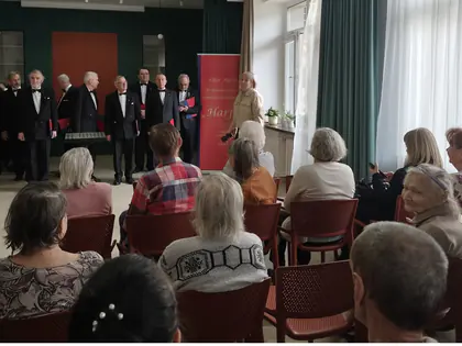 Męski chór w ciemnych garniturach śpiewa przed mieszkańcami siedzącymi w sali