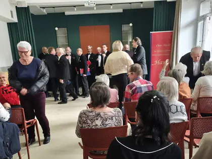 Chór męski przed zieloną ścianą, na pierwszym planie kobieta odwrócona do słuchaczy