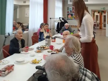 Mieszkańcy przy stole z kubkami, kobieta podaje szarlotkę