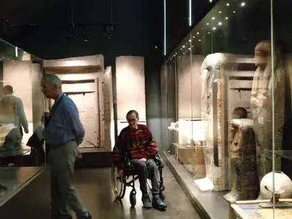 Ciemna sala muzeum z gablotami i sarkofagami, przechodzi mężczyzna, obok siedzi osoba na wózku