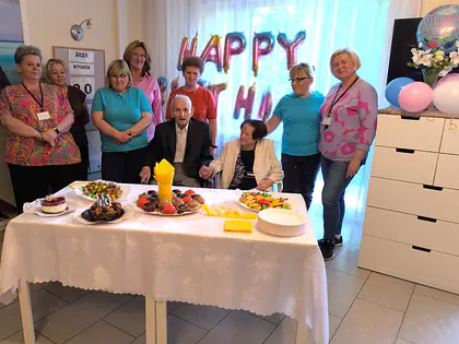 Pracownice i jubilat z żoną przy stole z ciastami i owocami, w tle balony z napisem Happy Birthday