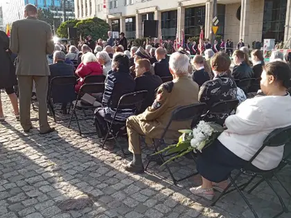 Uczestnicy uroczystości siedzą na krzesłach na skwerze przed budynkami