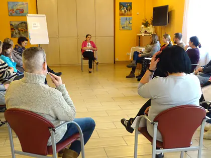 Uczestnicy spotkania siedzą w kole na krzesłach, w środku prowadząca