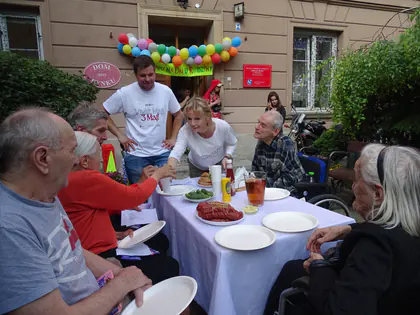 Mieszkańcy i goście przy stole z poczęstunkiem przed budynkiem ozdobionym balonami