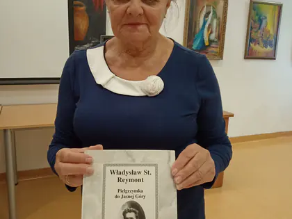 Kobieta trzyma broszurę Władysław St. Reymont, Pielgrzymka do Jasnej Góry, w sali z obrazami