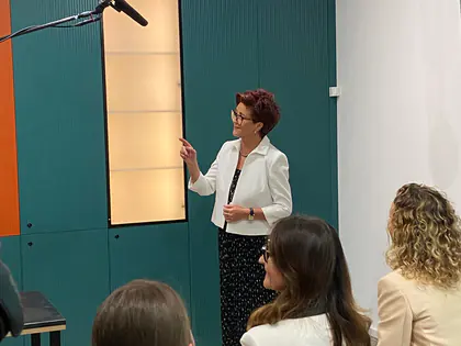 Kobieta w białym żakiecie przemawia do gości, przed nią siedzą słuchacze