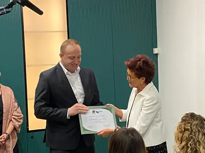 Mężczyzna w ciemnej marynarce odbiera dyplom od kobiety w białym żakiecie