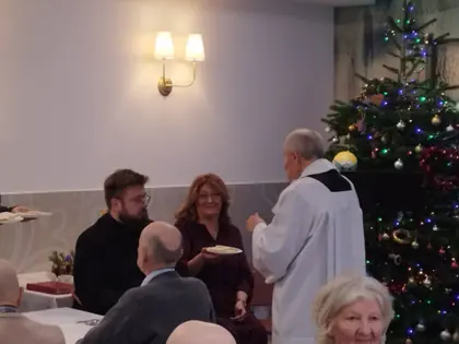 Ksiądz w komży dzieli się opłatkiem z uczestnikami wigilii, obok stoi kobieta z talerzem opłatków